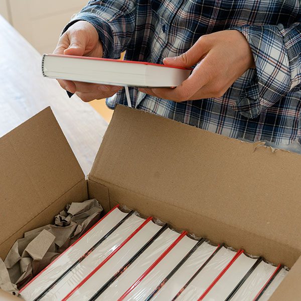 Person in a plaid shirt unboxing a shipment of books, holding one book while others remain stacked in the box.